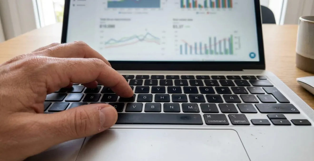 Primer plano de una mano escribiendo en el teclado de un ordenador portátil moderno con gráficos inmobiliarios en la pantalla de fondo