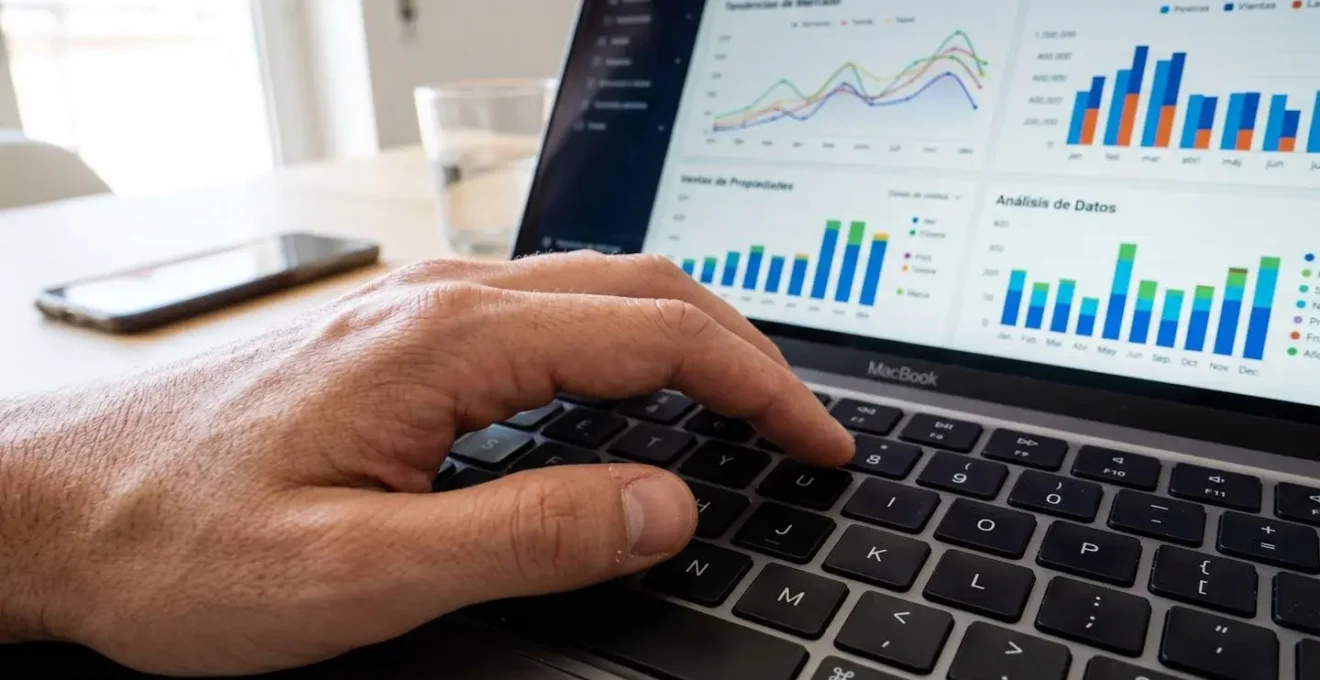 Primer plano de una mano escribiendo en el teclado de un ordenador portátil moderno con gráficos inmobiliarios en la pantalla de fondo